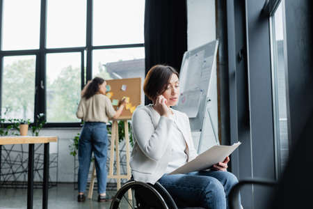 businesswoman in wheelchair talking on mobile phone while blurred african american colleague working...