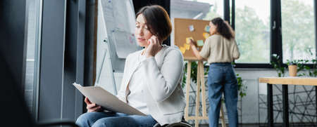 businesswoman with disability looking at documents and talking on smartphone near blurred african...