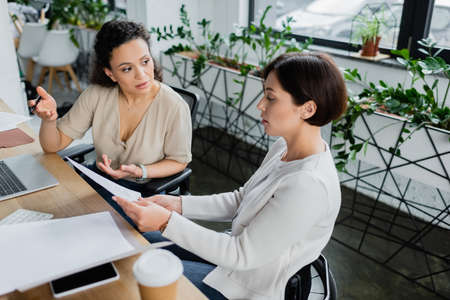 businesswoman with disability showing document to african american colleague during  conversation in officeの写真素材