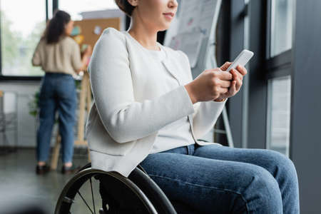 cropped view of businesswoman in wheelchair messaging on mobile phone near african american colleague on blurred backgroundの写真素材