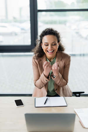 successful african american businesswoman showing win gesture near blurred laptop in officeの写真素材