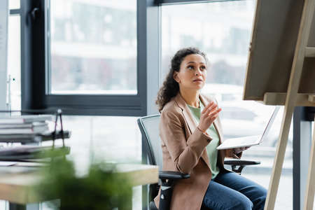 african american businesswoman sitting with laptop and pointing at note board in officeの写真素材