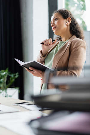 thoughtful african american businesswoman standing with notebook and pen at workplace in officeの写真素材