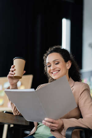 cheerful african american businesswoman holding coffee to go while working with documents in officeの写真素材