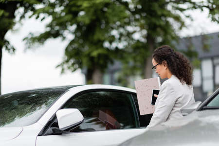 african american businesswoman with paper folder and mobile phone near car on parkingの写真素材