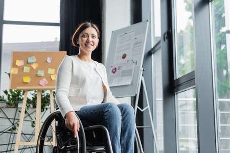 cheerful businesswoman in wheelchair looking at camera near blurred infographics in office
