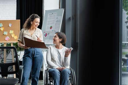 happy businesswoman in wheelchair talking to african american colleague near blurred flip chart in officeの写真素材