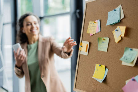 blurred african american businesswoman with smartphone pointing at corkboard with paper notesの写真素材