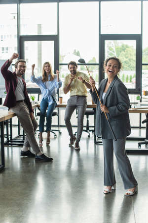 Excited african american businesswoman holding golf club near blurred excited colleagues in officeの写真素材