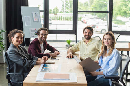 Positive multicultural business people looking at camera near papers and calculator in officeの写真素材