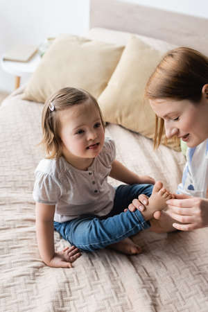 Smiling mother touching feet of baby daughter with down syndrome in bedroomの写真素材