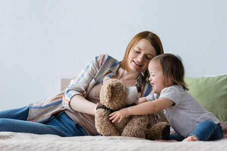 Smiling woman playing with daughter with down syndrome and soft toy on bedの写真素材