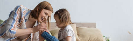 Smiling mother touching leg of child with down syndrome on bed, bannerの写真素材