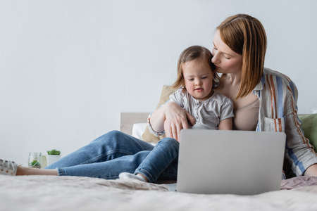 Mother kissing toddler daughter with down syndrome near laptop in bedroomの写真素材