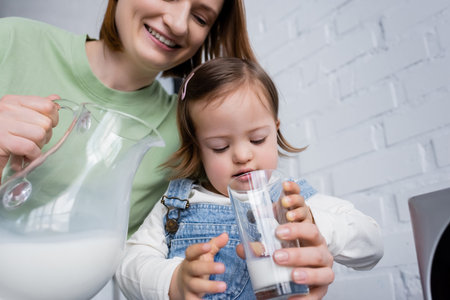 Smiling woman holding jug with milk near daughter with down syndrome in kitchenの写真素材