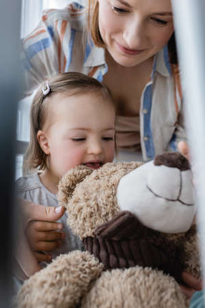 Cheerful kid with down syndrome looking at teddy bear near mother at homeの写真素材