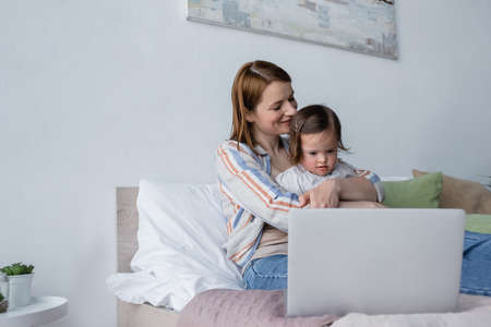 Happy mom hugging toddler daughter near laptop on bedの写真素材