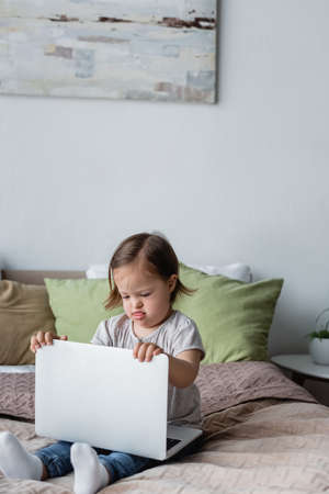 Child with down syndrome holding laptop on bed at home
