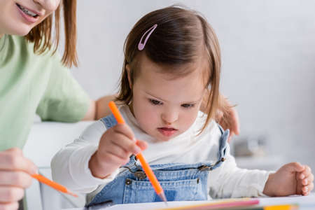Smiling woman holding color pencil near daughter with down syndrome at homeの写真素材