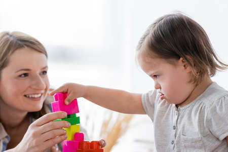 Concentrated kid with down syndrome playing building blocks near blurred smiling mother at home