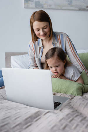 Smiling parent freelancer looking at daughter with down syndrome near laptop on bedの写真素材