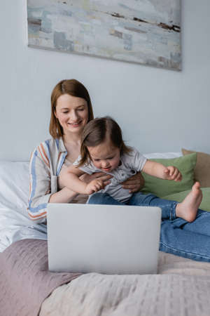 Smiling woman holding child with down syndrome near laptop in bedroomの写真素材