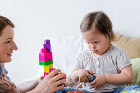 Focused child with down syndrome looking at building blocks near smiling mom in bedroomの写真素材