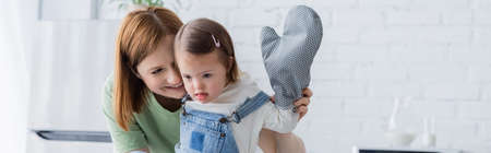 Happy mother looking at child with down syndrome and baking glove in kitchen, banner