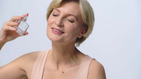 happy mature woman with closed eyes holding perfume isolated on white