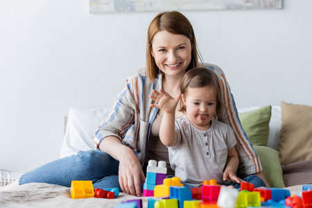 Woman and child with down syndrome waving hand at camera near building blocks on bedの写真素材