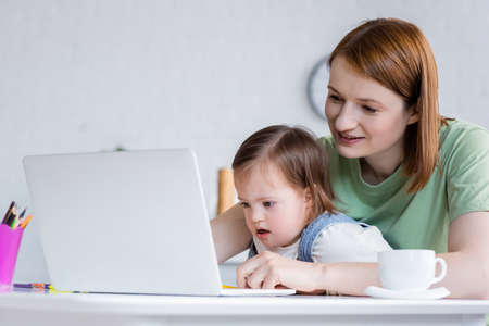 Smiling freelancer using laptop near daughter with down syndrome and coffee at homeの写真素材