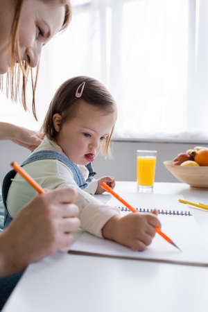 Child with down syndrome drawing on paper near mother and orange juice in kitchenの写真素材