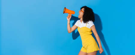 energized african american woman in trendy jumpsuit screaming in megaphone on blue, bannerの写真素材