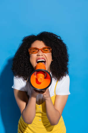 thrilled african american woman in orange eyeglasses screaming in megaphone on blueの写真素材