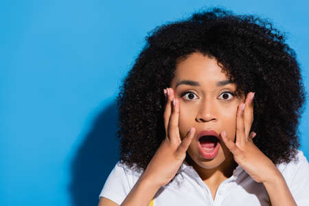 shocked african american woman touching face while looking at camera on blue