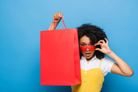 african american woman touching stylish eyeglasses while showing coral shopping bag on blueの写真素材
