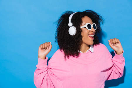 cheerful african american woman in stylish sunglasses listening music in headphones on blueの写真素材