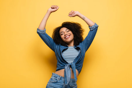 joyful african american woman in denim shirt dancing with closed eyes and raised hands on yellowの写真素材