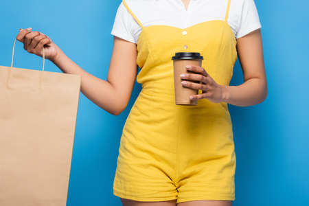 partial view of african american woman in yellow jumpsuit holding shopping bag and paper cup on blueの写真素材
