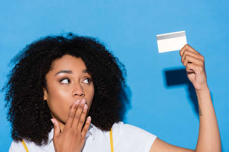 amazed african american woman covering mouth with hand while holding credit card on blue