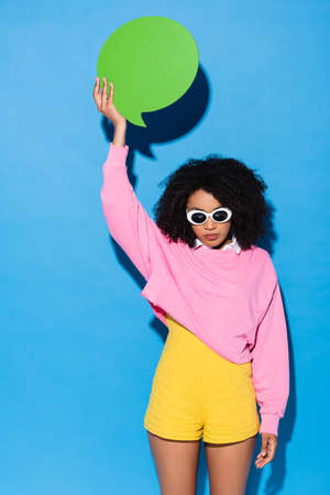 african american woman in stylish sunglasses holding empty thought bubble in raised hand on blue