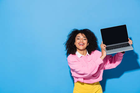 cheerful african american woman holding laptop with blank screen on blueの写真素材