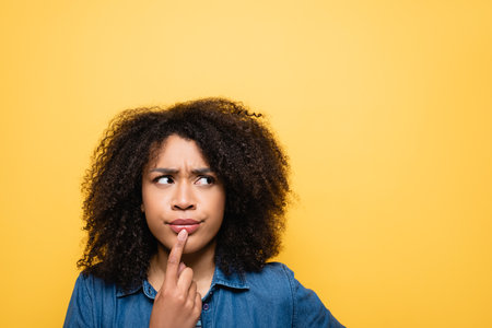 thoughtful african american woman touching lips and looking away isolated on yellowの写真素材