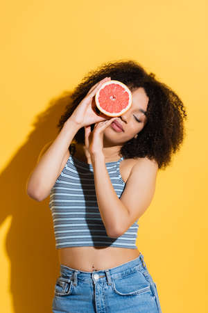 young african american woman in striped tank top obscuring face with half of ripe grapefruit on yellowの写真素材
