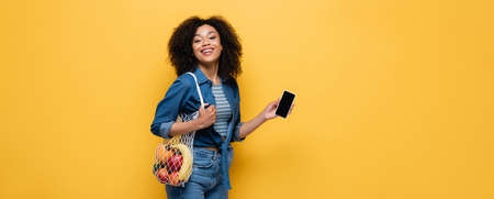 happy african american woman holding smartphone and string bag with fruits isolated on yellow,...