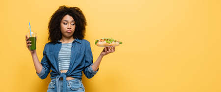 confused african american woman looking at hot dog while holding fresh smoothie on yellow, bannerの写真素材