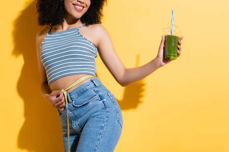 partial view of pleased african american woman measuring waist while holding fresh smoothie on...