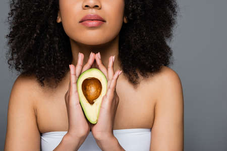 cropped view of african american woman in white top holding half of avocado isolated on greyの写真素材