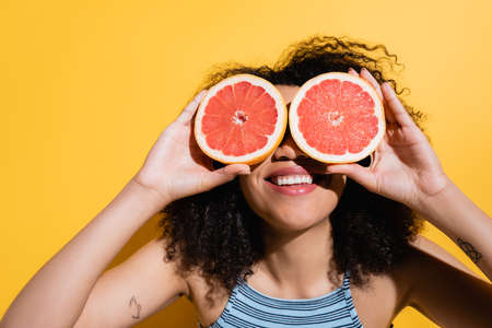 cheerful african american woman obscuring face with halves of juicy grapefruit on yellowの写真素材