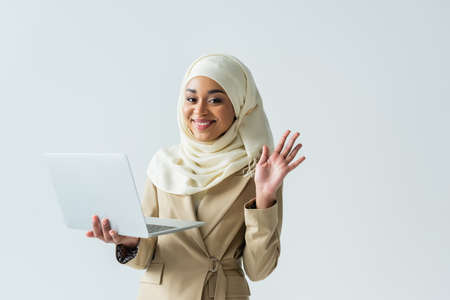 cheerful muslim woman in hijab and beige suit holding laptop and waving hand isolated on greyの写真素材